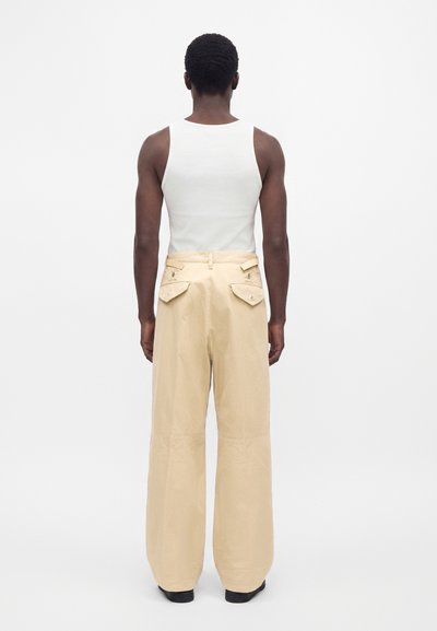 Personne de dos portant un top blanc sans manches et un pantalon beige ample avec poches à rabat à l'arrière sur un fond blanc uni.