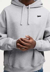 Harmaa huppari, valmistettu pehmeästä kankaasta, jossa kengurun tasku, kiristysnauhallinen huppu ja pieni musta logo rinnassa.