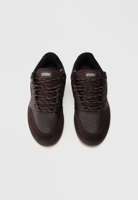 Brūni sporta apavi ar gludas un zamšādas tekstūras sajaukumu, perforētiem akcentiem un melnām šņorēm. Uz mēlītes redzams Etnies logotips.