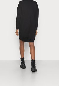 Černé oversize šaty ve stylu mikiny s dlouhými rukávy, žebrovanými manžetami a zakřiveným lemem, doplněné robustními černými botami na šněrování s knoflíkovými detaily.