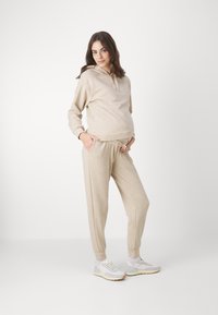 Beige huvtröja och matchande joggers, mjuk textur, avslappnad passform, framfickor, dragsko i midjan, i kombination med ljusgrå sneakers.