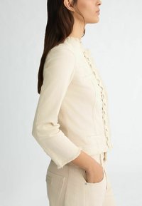 Giacca cropped color crema con bordi sfilacciati, caratterizzata da un davanti in pizzo texturizzato con laccatura e maniche a tre quarti, abbinata a pantaloni coordinati.