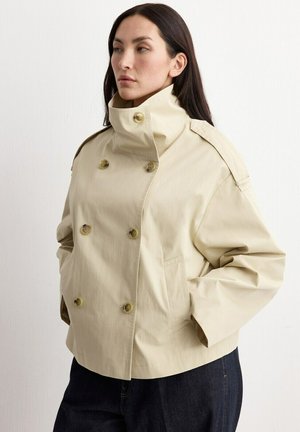 Kobieta ubrana w beżową dwurzędową kurtkę z wysokim kołnierzem i bocznymi kieszeniami, zestawioną z ciemnymi spodniami na jednolitym tle.