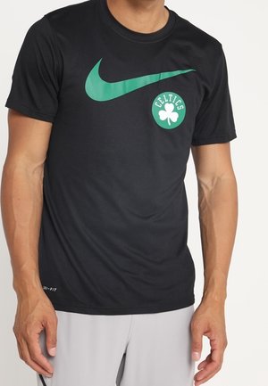 Mann trägt ein schwarzes Nike-T-Shirt mit grünem Celtics-Logo und Swoosh, kombiniert mit hellgrauen Shorts.