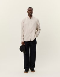 Ανοιχτό μπεζ πουκάμισο με μακριά μανίκια, με επαγγελματικό γιακά και κουμπί, συνδυασμένο με σκούρες φαρδιές παντελόνες και μαύρα παπούτσια, κρατώντας ένα μαύρο καπέλο.