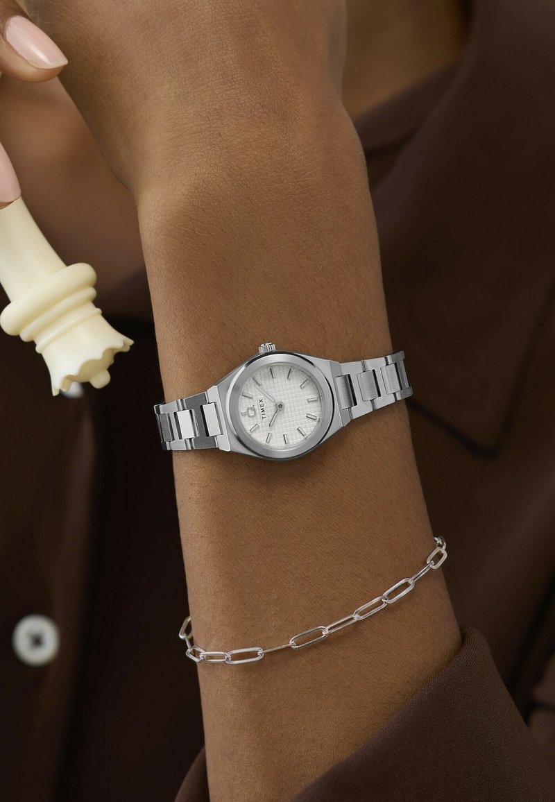 Κα Wrist με ασημένιο ρολόι Timex και λεπτό ασημένιο βραχιόλι αλυσίδα, χέρι που κρατά λευκό πιόνι της βασίλισσας σκακιού με φόντο καφέ πουκάμισο.