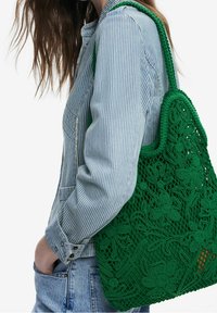 Vihreä virkattu laukku kukkakuviolla, jossa on jämäkkä muoto ja paksu teksturoitu olkahihna, yhdistettynä siniraidalliseen paitaan.