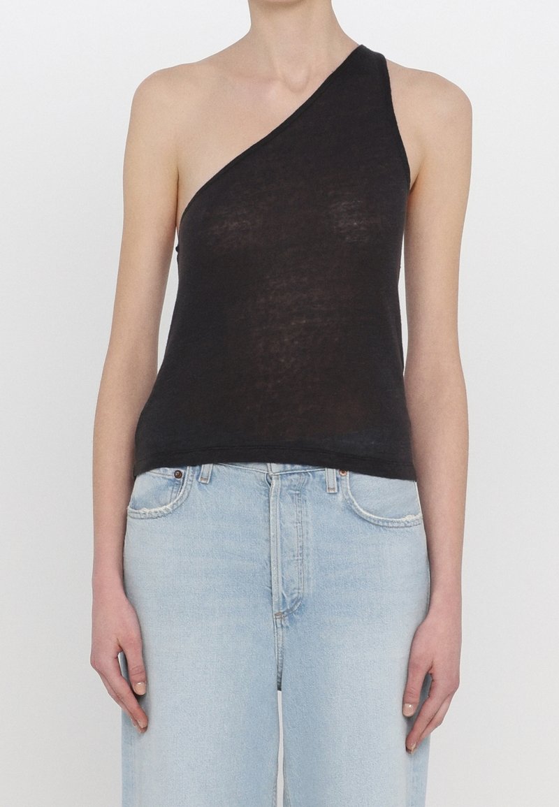 Personne portant un haut noir asymétrique sans manches et un jean taille haute bleu clair, debout avec les bras détendus sur les côtés.