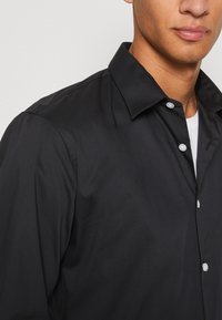 Camicia nera con bottoni a pressione, colletto strutturato, tessuto liscio e bottoni perlati; visibile parzialmente su un modello su uno sfondo chiaro.