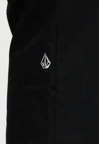 Μαύρο ύφασμα με ένα μικρό λευκό κεντητό γεωμετρικό λογότυπο που μοιάζει με σχήμα διαμαντιού κοντά στο κέντρο.