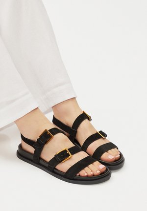 Piedi con sandali neri con tre cinturini spessi e fibbie dorate, abbinati a pantaloni bianchi su uno sfondo bianco uniforme.