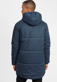 Geacă căptușită de culoare bleu marin cu glugă, având oつ quilting orizontală, mâneci lungi și un spate ușor prelungit pentru o acoperire suplimentară.