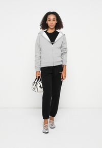 Grauer Zip-Hoodie mit weißem Innenfutter, kombiniert mit schwarzen Jogginghosen. Silberne Ballerinas mit Riemen und einer kleinen weißen Handtasche.