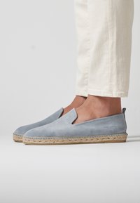 Zapatos de ante de color azul claro, tipo slip-on, con suela de yute, que presentan una punta cerrada y laterales recortados, mostrando un sutil logotipo en el talón.