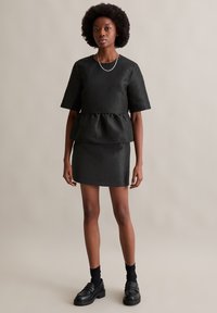 Melnā pepluma krekls un pieguļošas svārki, izgatavoti no strukturētas auduma. Modelis valkā melnus loaferus un zeķes, kā arī vienkāršus sudraba kakla rotājumus. Fons ir vienkāršs.