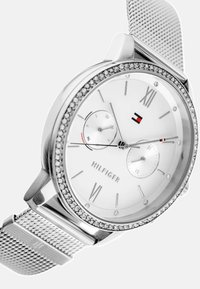 Orologio argento con quadrante bianco, numeri romani, due sotto-quadranti e un quadrante incastonato di cristalli. Presenta un cinturino a rete e il logo del marchio.
