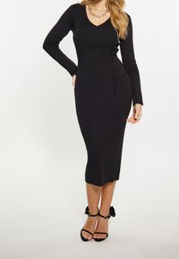 Robe midi noire côtelée avec manches longues et encolure en V, dotée d'un détail noué à la taille. Portée avec des talons noirs à lanières.