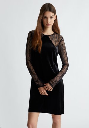 Abito in velluto nero con maniche lunghe in pizzo. Collo rotondo, silhouette aderente e dettagli in pizzo trasparente, creando un contrasto di texture e design.