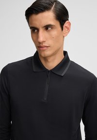 Polo shirt nero a maniche lunghe realizzato in tessuto testurizzato, con chiusura a zip frontale e rifinitura a contrasto del colletto. Design minimalista.