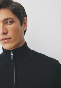 Pulover cu guler înalt, negru, cu șiret metalic, prezentând un model de tricot texturat și o croială ajustată.