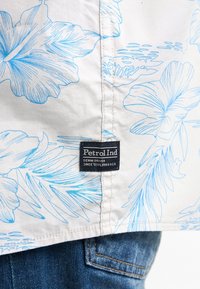 Camicia bianca con motivo floreale blu; presenta un'etichetta nera con la scritta "Petrol Ind" vicino all'orlo; jeans in denim visibili sotto.