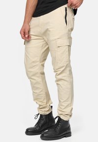 Pantaloni cargo beige con molte tasche, design affusolato e dettagli con cerniera. Indossati con stivali neri stringati; realizzati in tessuto resistente.