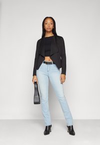 Svart cropped blazer med drapert front, kombinert med lyseblå rette jeans og svarte ankelstøvler. Holder en svart skulderveske.