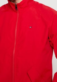 Röd zip-up jacka med stående krage, liten logotyp på bröstet och sned framlucka på högra sidan.