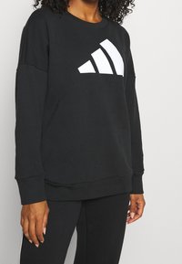 Sudadera negra con un gran logo de tres franjas blancas. Presenta un cuello redondo, mangas largas y un corte relajado. Textura de tela suave.