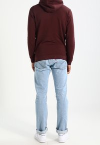 Felpa con cappuccio color bordeaux, jeans azzurri chiari, sneaker bianche. Attenzione al taglio casual con una consistenza morbida; cuciture a vista e dettagli di orlo risvoltato.