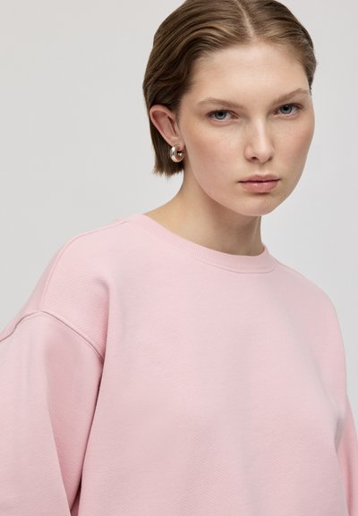 Felpa rosa chiaro con girocollo, spalle scese e tessuto struttura. Modella accessoriata con orecchini a cerchio in argento.