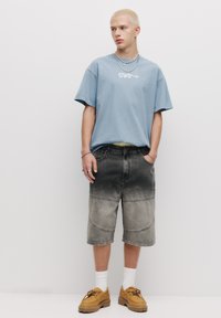Tricou oversized de culoare albastru deschis, cu text brodat, asortat cu șorturi din denim gri de lungime până la genunchi, cu detalii din panouri. Pantofi călcați maro cu șireturi.