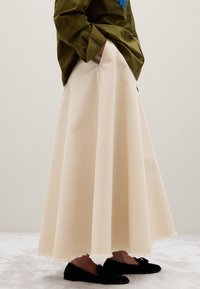 Personne portant une longue jupe beige avec un ourlet effiloché, une chemise vert olive avec une broderie bleue, les mains dans les poches, et des mocassins noirs.