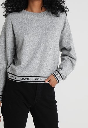 Person iført en lysegrå langærmet sweatshirt med Levi's-logo på ærmekanter og kant, kombineret med sorte bukser.