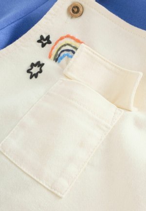 Crèmekleurige stoffen zak met gestikte regenboog- en sterontwerpen, bevestigd aan een blauw kledingstuk. Knopdetail zichtbaar boven de zak.