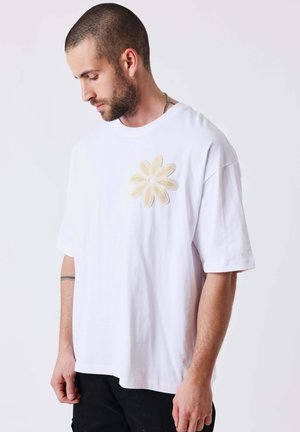 Man met kort haar en baard, draagt een wit oversized T-shirt met een beige madeliefapplicatie op de borst, kijkt naar beneden zijwaarts.