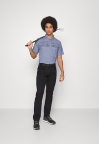 Golf-Polohemd in Hellblau mit gemusterten Brustakzenten, kombiniert mit schwarzen Hosen und schwarzen Sportschuhen. Männliches Model hält einen Golfschläger.