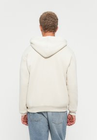 Persona con cabello rizado corto, vistiendo una sudadera sencilla de color blanco roto y jeans de color azul claro, de espaldas contra un fondo blanco.