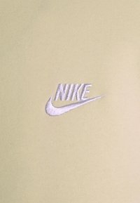 Světle zelená tkanina s hladkou strukturou, na které se nachází bílé vyšívané logo Nike se swoosh designem.