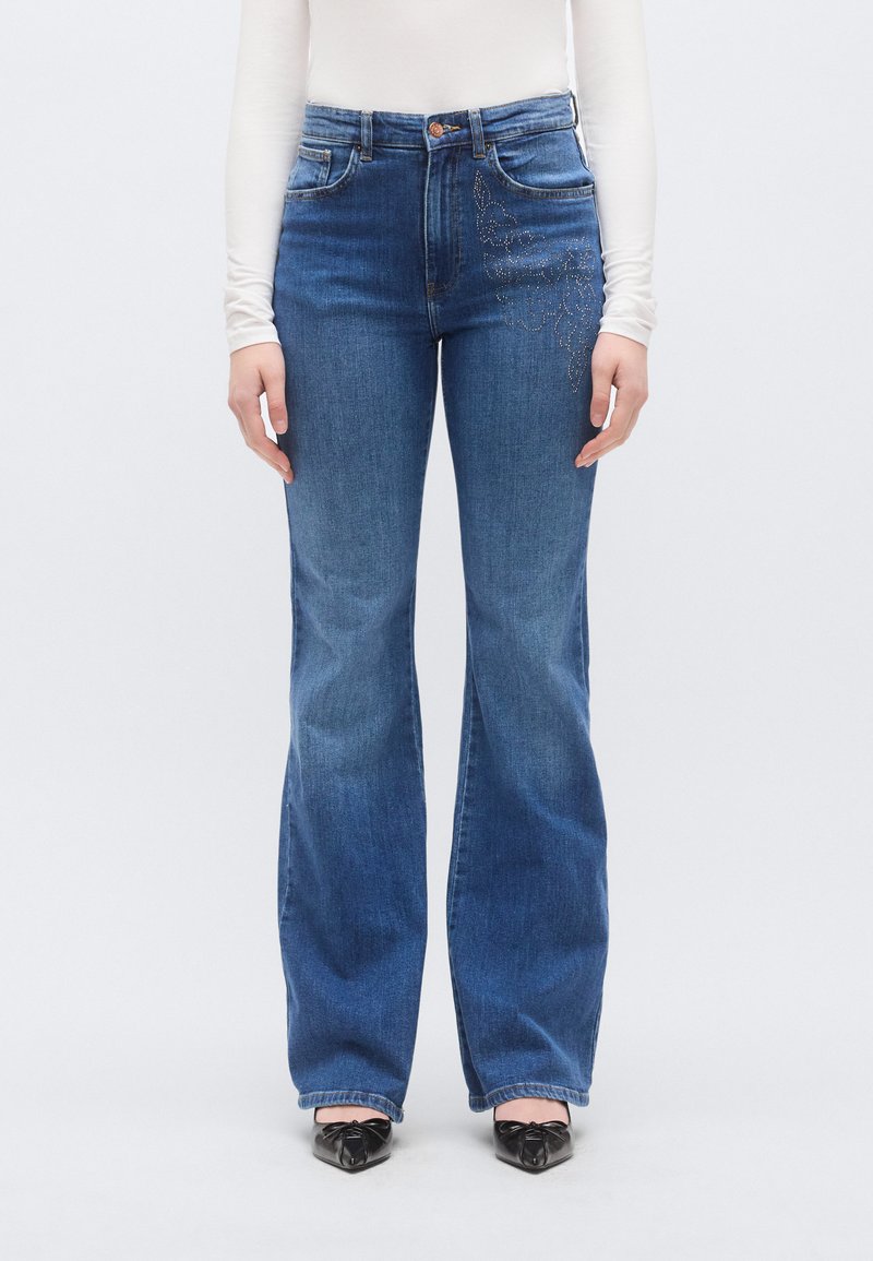 Τζιν με ψηλή μέση και καμπάνα σε μεσαίο μπλε ντένιμ, με διακοσμητικό λουλουδάτο μοτίβο από στρας στον αριστερό μηρό.