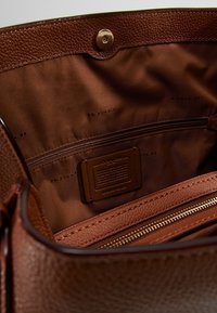 Interiér hnědé kožené kabelky s hladkým nylonovým podšívkou, obsahující zipovou kapsu a raženou značkovou etiketu. Viditelné jemné detaily textury.