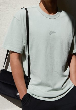 Personne portant un t-shirt Nike vert clair et un sac à bandoulière Nike noir, debout contre un mur clair texturé avec une main dans la poche.
