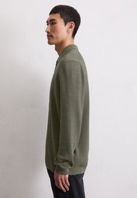 Polo a maniche lunghe, di colore verde, realizzato in tessuto testurizzato, con colletto e polsini a coste, abbinato a jeans scuri.