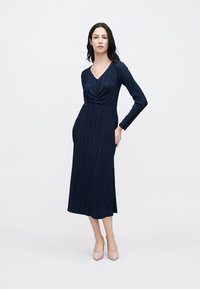 Tumši zila kleita ar garām piedurknēm, savāktā v-veida izgriezumā un vertikālu riboto tekstūru. Kleita sniedzas līdz vidusvaskam, saskaņota ar gaiši rozā augstpapēžu kurpēm.