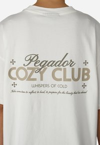 Balts kokvilnas t-krekls ar lielu tekstu bēšā krāsā. Uzraksts "Pegdor COZY CLUB WHISPERS OF COLD" ar dekoratīviem akcentiem zem teksta.