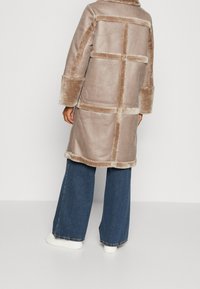 Beige shearling coat with a grid design, fur accents, wide cuffs, and a rounded collar, paired with blue wide-leg jeans and white shoes.