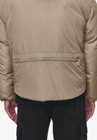 Beige jakke med fyll som har en glatt, blank overflate, med en glidelåslomme på brystet og elastiske mansjetter. Bakvisning viser en buet søm.