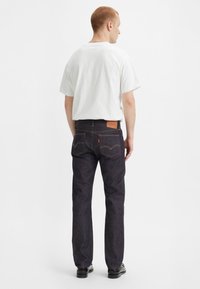 Jeans de denim azul oscuro con corte recto, parche de cuero en la parte trasera y costuras amarillas; combinados con una camiseta blanca.