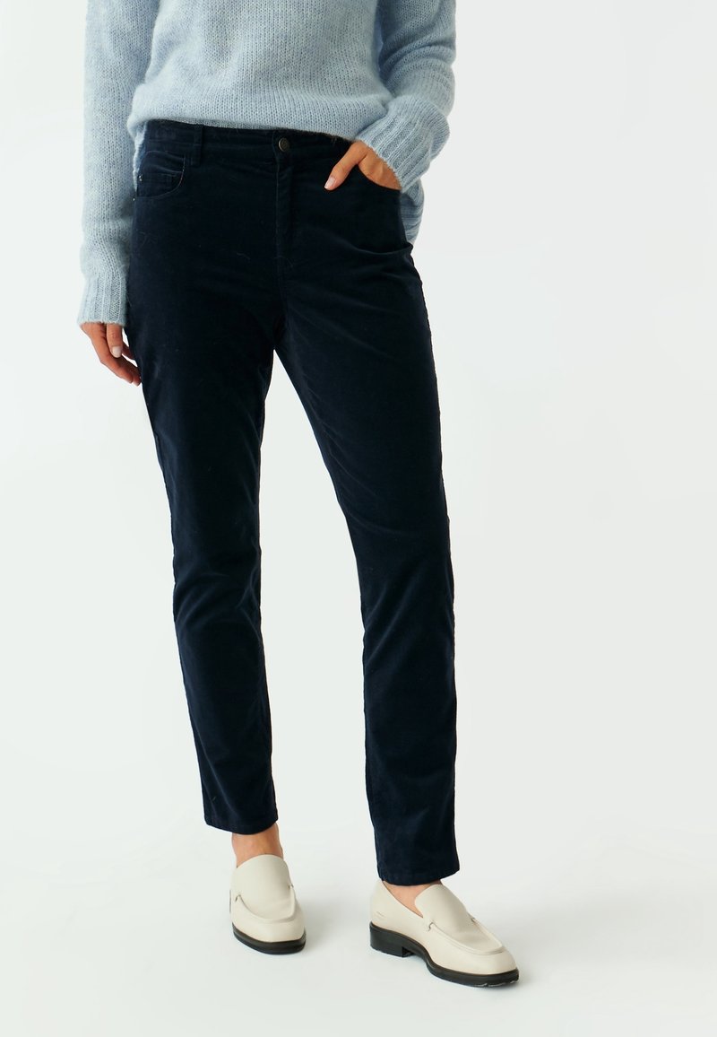 Personne portant un pantalon en velours côtelé noir, un pull bleu clair, des mocassins couleur crème, debout avec une main dans la poche, sur un fond blanc.