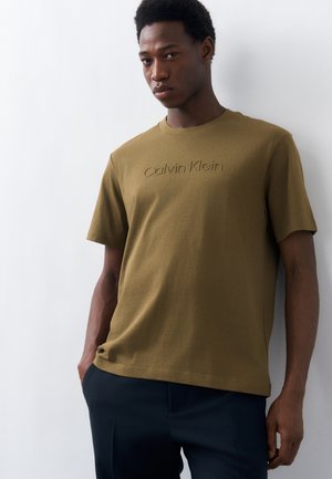 Νέος άνδρας που φοράει ελιάς πράσινο t-shirt Calvin Klein και σκούρο παντελόνι, στέκεται μπροστά σε λευκό τοίχο με τα χέρια στις τσέπες.